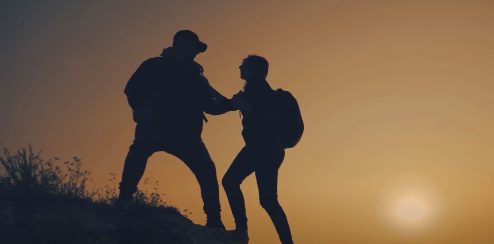 Silhouette of a guy and a girl helping each other while climbing an elevated ground