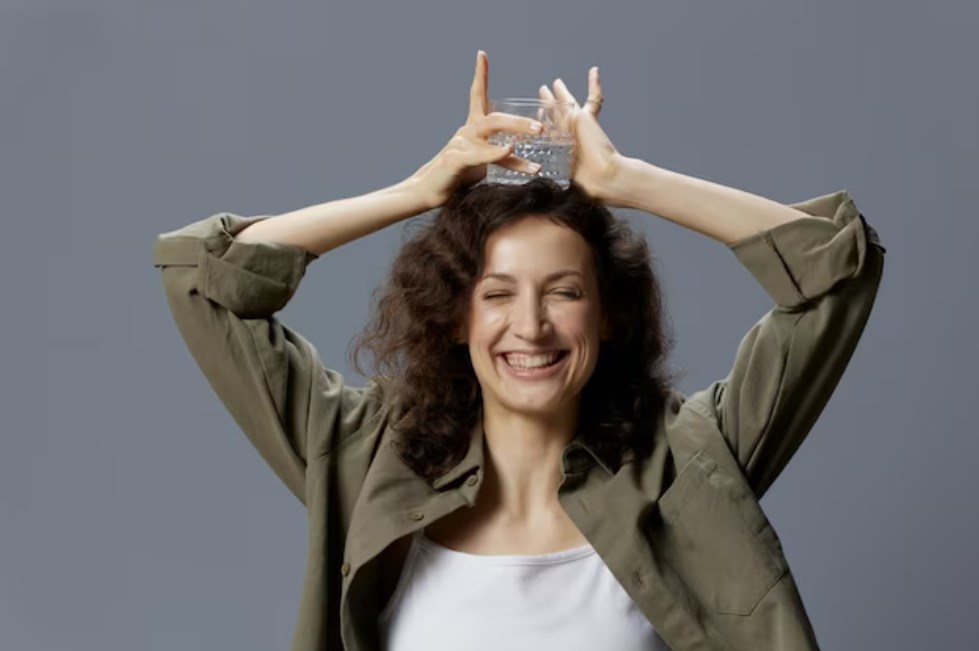 a smiling curly woman in casual khaki-green shirt holding a glass of water overhead