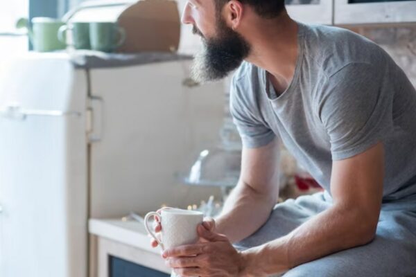 a man in a grey t-shirt sitting with a cup on a kitchen counter and looking sideways