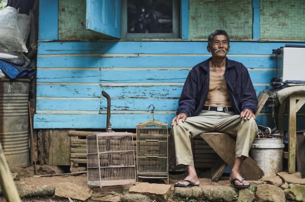 an elderly Asian man sitting and smiling outside a traditional-wooden house