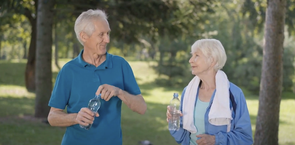 Photo of an elderly man and a woman standing, holding water bottles, looking at each other and smiling