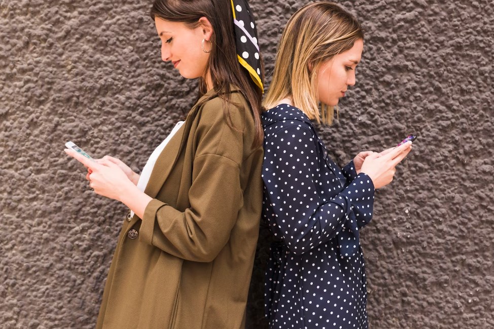 two young female friends standing back-to-back and texting on their phones