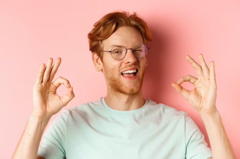 a ginger guy in glasses winking and showing okay signs in a pink background