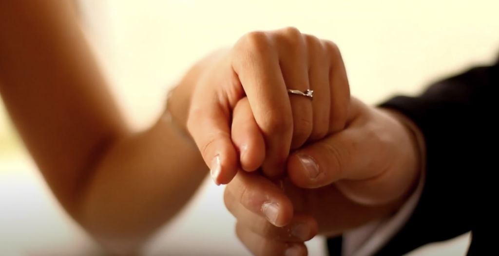 Photo of two hands holding each other, with the female's hand adorned with a ring