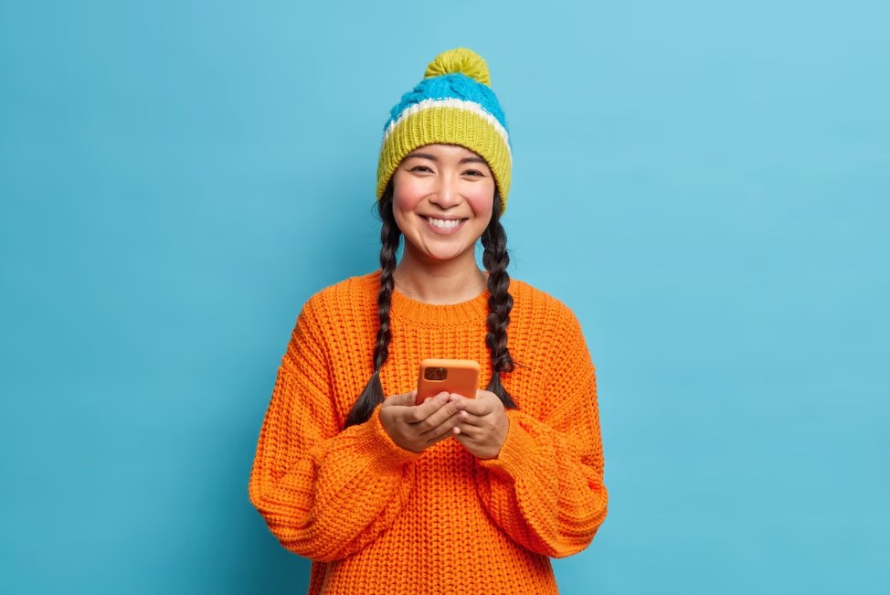 a pretty smiling Asian girl with two pigtails wearing a green-and-blue hat and an orange sweater
