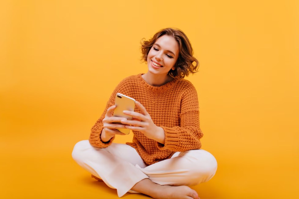 a positive barefooted girl sitting in a yellow background with her phone