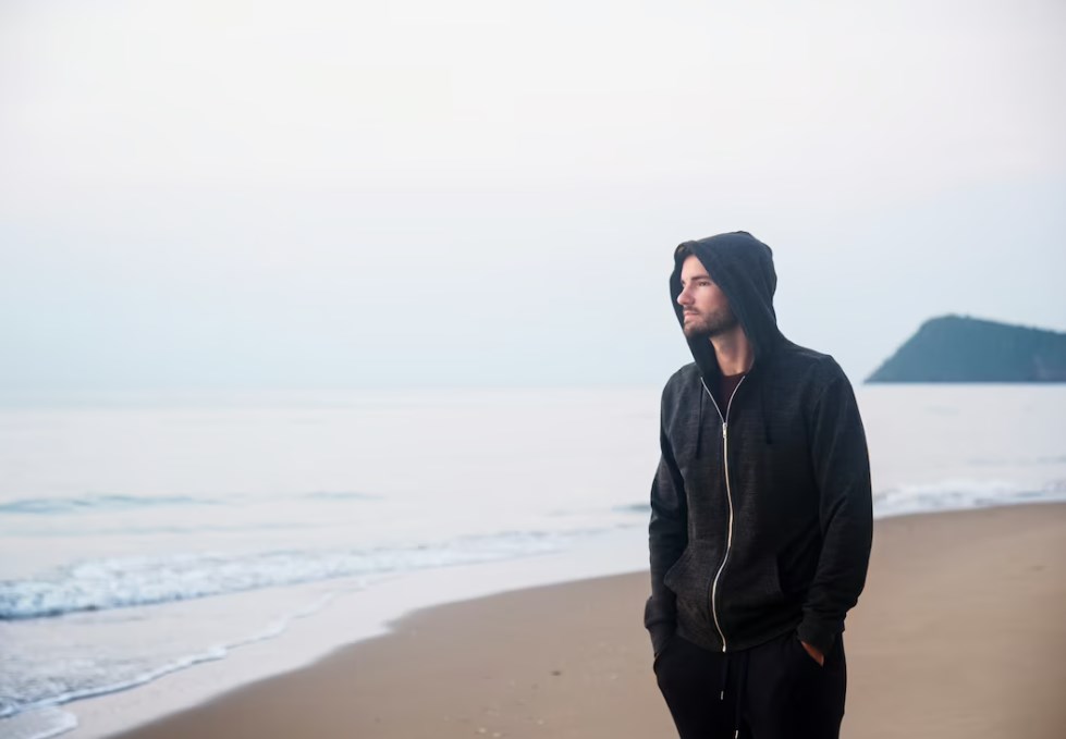 a man in a black zip-up hoodie walking on the beach and looking at the water