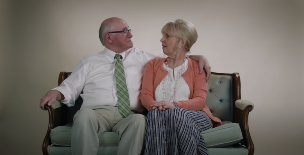 Photo of an old man and an old woman engaged in conversation, with the man's arm around the woman's shoulder